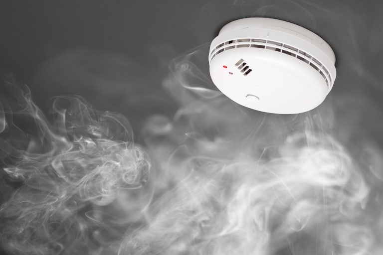 A ceiling-mounted smoke detector with a red light is surrounded by wisps of smoke against a dark background.