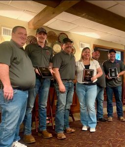 Six people stand indoors, smiling and holding plaques or awards. They are dressed casually, mostly in jeans and gray shirts, and are posing together in front of a wall with decorative plates.