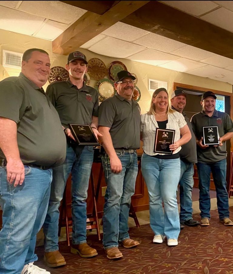Six people stand indoors, smiling and holding plaques or awards. They are dressed casually, mostly in jeans and gray shirts, and are posing together in front of a wall with decorative plates.