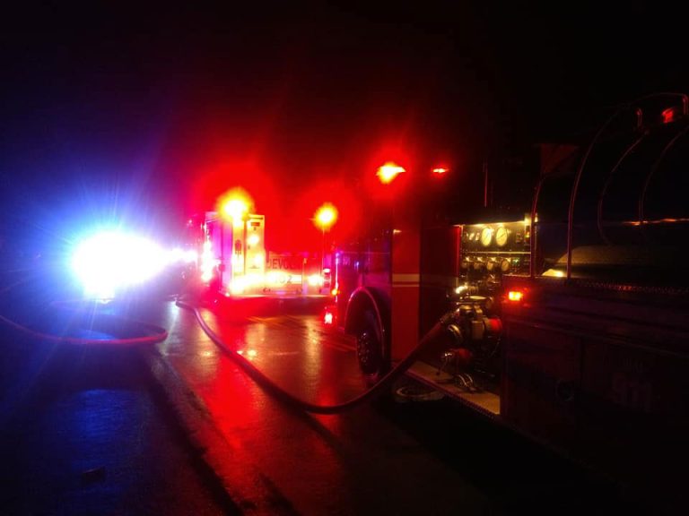 A fire truck with bright red emergency lights is parked on a dark road at night, with hoses attached and another vehicle with blue lights visible in the background.