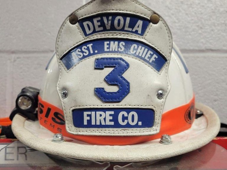 A white firefighter helmet with blue and white labels reading "DEVOLA," "SST. EMS CHIEF," a large number "3," and "FIRE CO." An orange band and a flashlight are attached to the helmet.