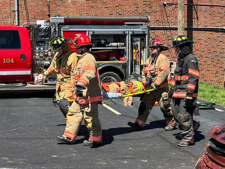 Four firefighters in full gear carry a person on a stretcher in a parking lot, with a fire truck and brick building in the background. The scene appears to be part of a rescue or training exercise.