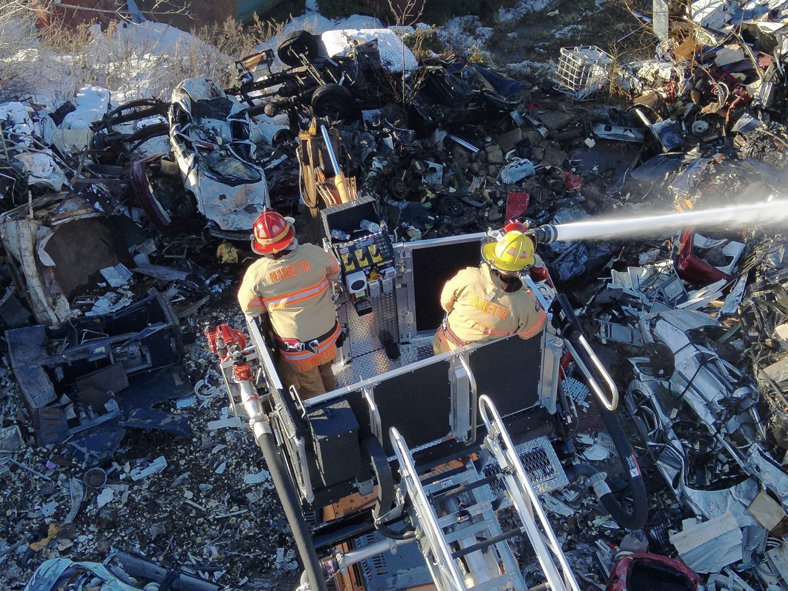 Two firefighters wearing helmets and uniforms stand on an elevated ladder truck, spraying water onto a pile of wrecked vehicles and debris at a junkyard during daylight.