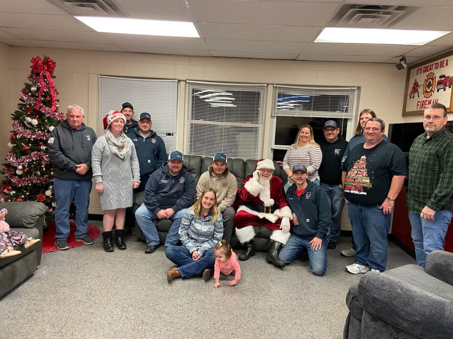 A group of people poses indoors with a decorated Christmas tree and Santa Claus; some wear festive hats. A young child is sitting on the floor in front of Santa, and everyone is smiling at the camera.