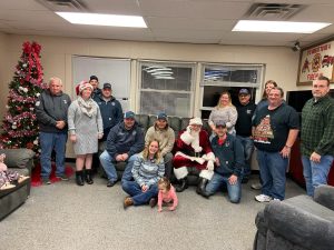 A group of people poses indoors with a decorated Christmas tree and Santa Claus; some wear festive hats. A young child is sitting on the floor in front of Santa, and everyone is smiling at the camera.