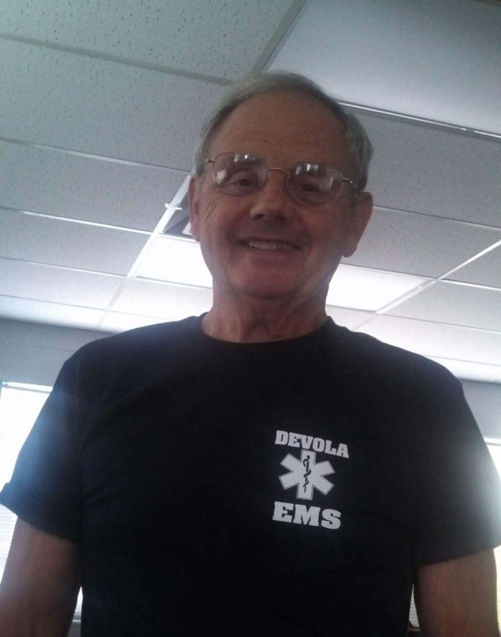 An older man with glasses smiles at the camera while wearing a black "Devola EMS" t-shirt. He is indoors with ceiling tiles and windows in the background.