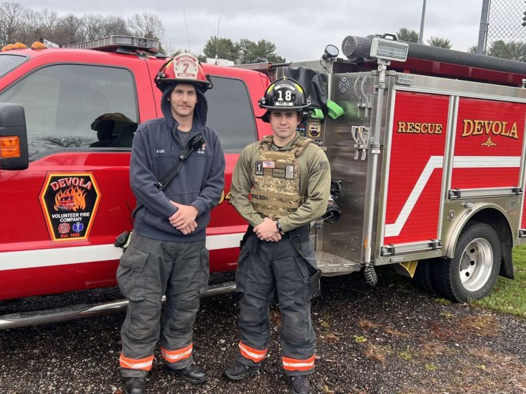 Two firefighters in uniform and helmets stand in front of a red Devola Volunteer Fire Department rescue truck on a cloudy day.