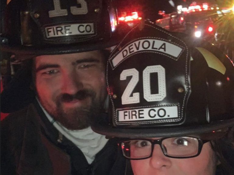 Two firefighters wearing black helmets labeled "13 FIRE CO." and "20 DEVOLA FIRE CO." pose for a selfie at night, with emergency vehicle lights flashing in the background.