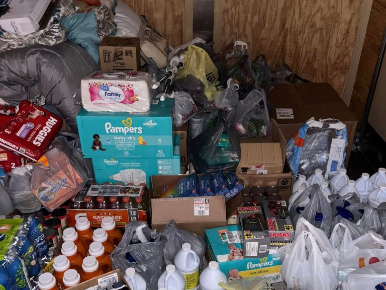 A large collection of donated supplies, including diapers, bottled drinks, canned food, household goods, and bags filled with various items, stacked and piled inside a room with wooden walls.