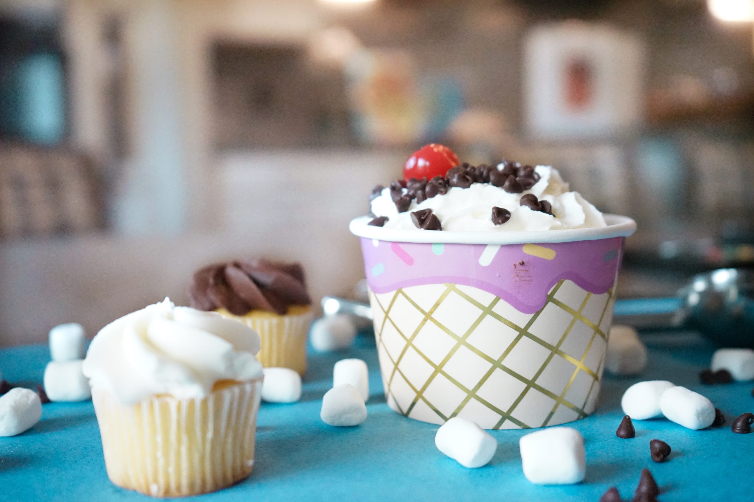 A close-up of a dessert cup filled with whipped cream, chocolate chips, and a cherry, surrounded by mini marshmallows, chocolate chips, and two frosted cupcakes on a blue surface. The background is blurred.