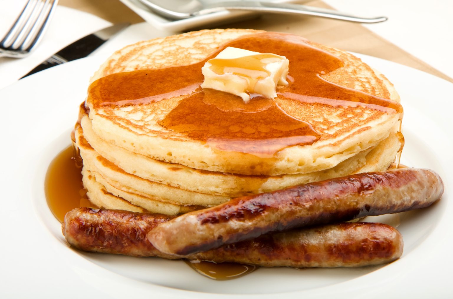 A stack of pancakes topped with a pat of butter and syrup, served with two sausage links on a white plate. A fork, knife, and napkin are in the background.