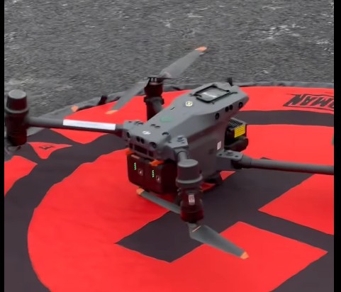 A gray quadcopter drone with four propellers is resting on a red and black circular landing pad placed on a rough, dark surface. The drone has visible sensors and lights on its body.