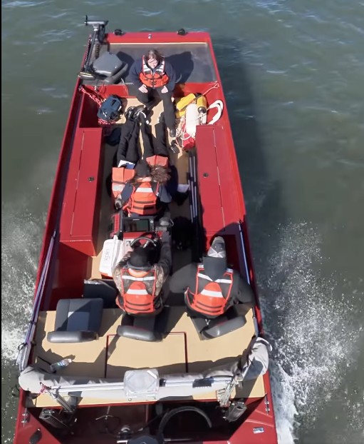 Aerial view of four people wearing orange life vests sitting in a red motorboat traveling on water. The boat creates a wake behind it as it moves forward.