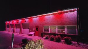 A brick fire station building is lit with red lights at night. The building has several garage doors on the left and large windows on the right, with a bench and some bushes in front.