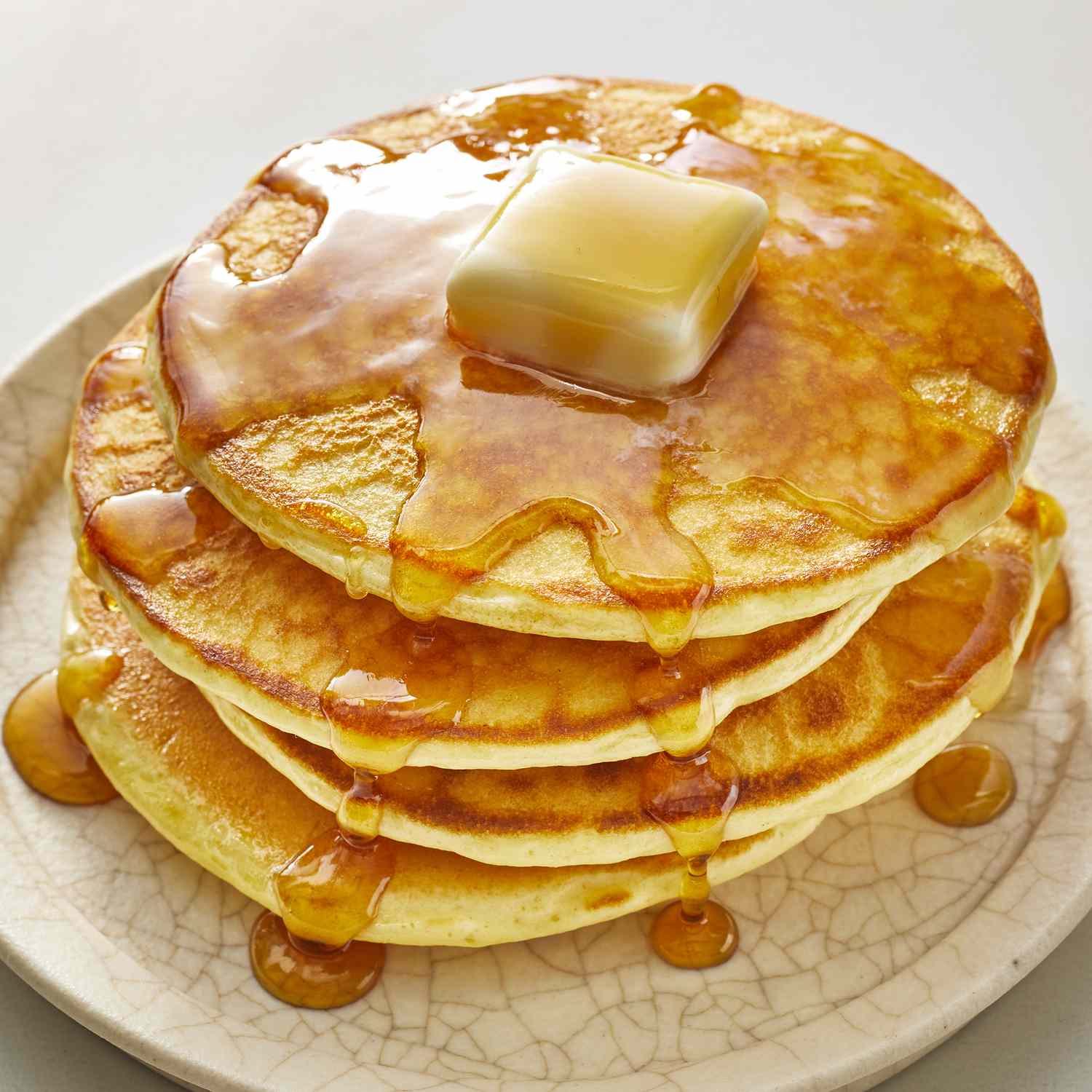 A stack of three golden pancakes topped with a pat of melting butter and drizzled with syrup on a light-colored plate.
