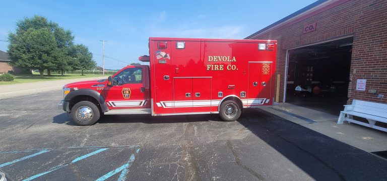 A bright red Devola Fire Co. emergency vehicle is parked outside a brick fire station on a sunny day, with the garage door open and green trees visible in the background.