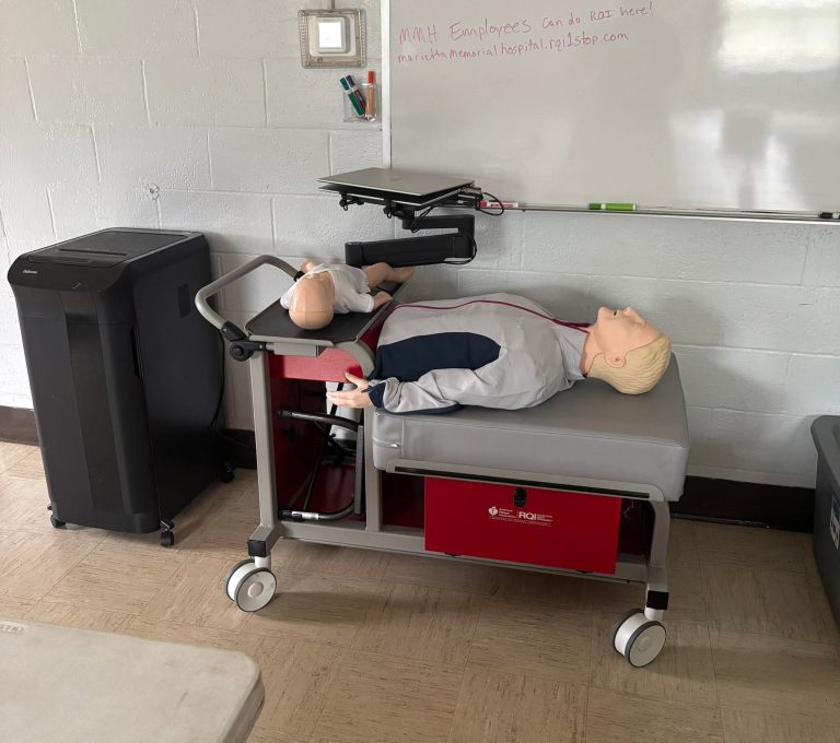 A medical training mannequin lies on a wheeled hospital bed next to a black machine, in a room with a whiteboard on the wall. The mannequin is positioned as if for practice or demonstration purposes.
