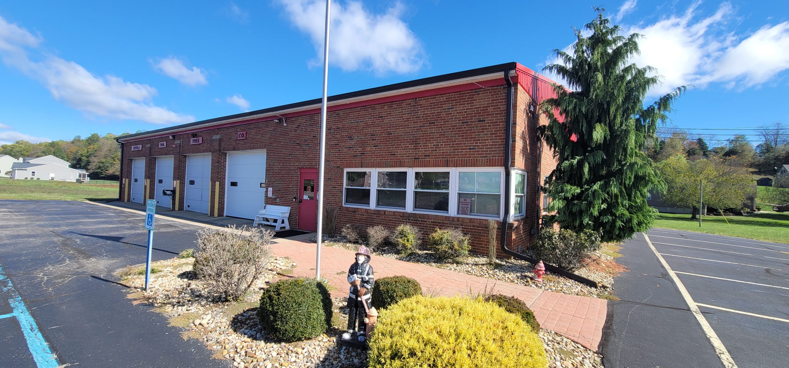 A brick fire station with three white garage doors and a red door sits under a blue sky. Shrubs and a firefighter statue are in front, with a flagpole at the center and a parking lot surrounding the building.