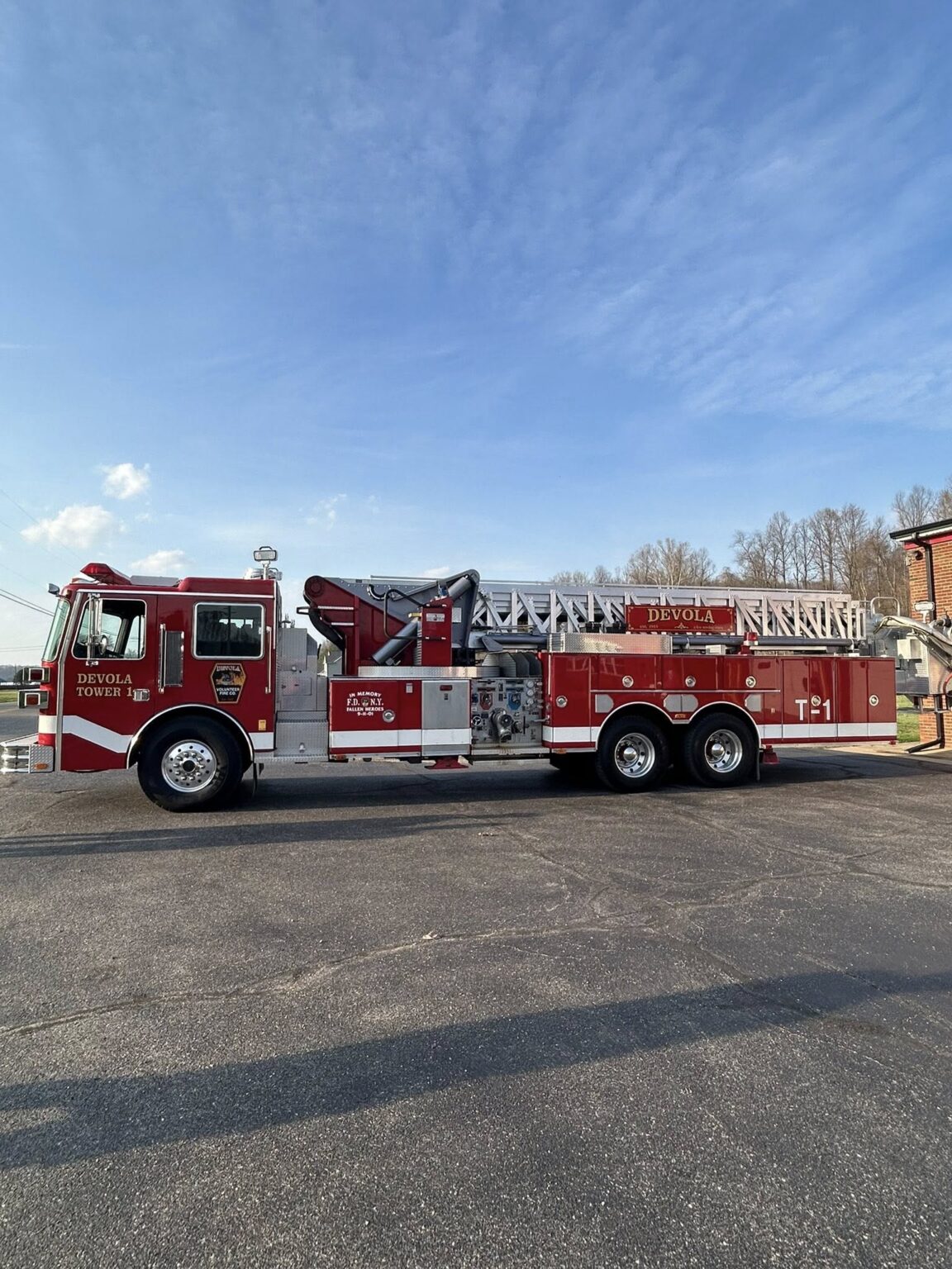 A red fire truck with an extended ladder on top is parked on a paved surface under a blue sky. The truck has "Devola Tower 1" and "T-1" written on the side. Trees and a building are in the background.