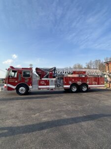 A red fire truck with an extended ladder on top is parked on a paved surface under a blue sky. The truck has "Devola Tower 1" and "T-1" written on the side. Trees and a building are in the background.
